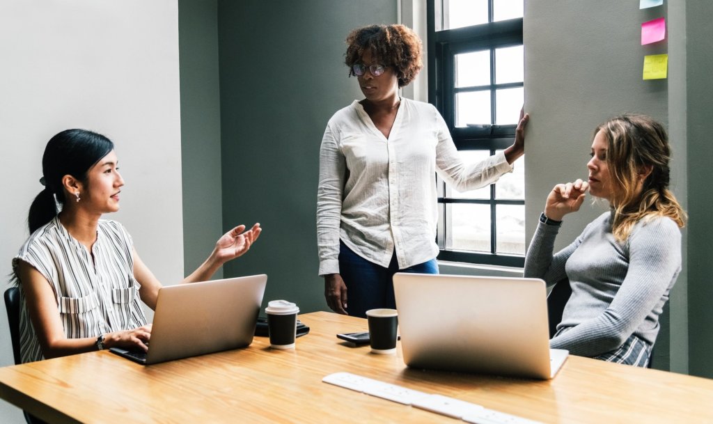 three people in a meeting. Two sitting with laptops and one standing